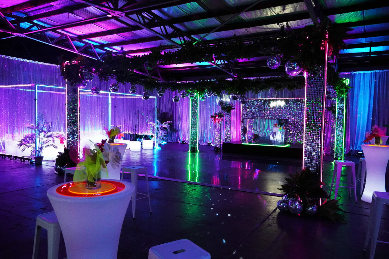 Room shot of neon disco party theme featuring illuminated furniture, bar stools, greenery, dance floor, neon lights, and sequin panels