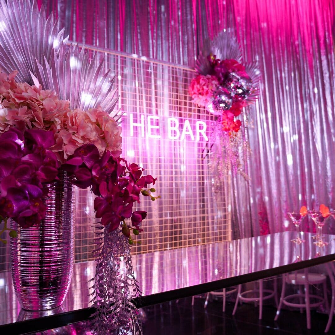 the bar at a pink disco dinner party setup. Area features a pink acrylic bar, florals, a mesh backdrop, and a pink neon 'the bar' sign