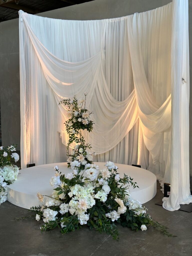 2025 wedding décor trends Gathered dramatic drape wedding ceremony backdrop with white stage and lush white and green florals
